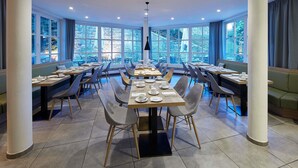 A spacious dining area with multiple tables and chairs, large windows offering a view of the outdoors, and a modern ceiling design with recessed lighting. - ibis Styles Neckarsulm  (Neckarsulm)