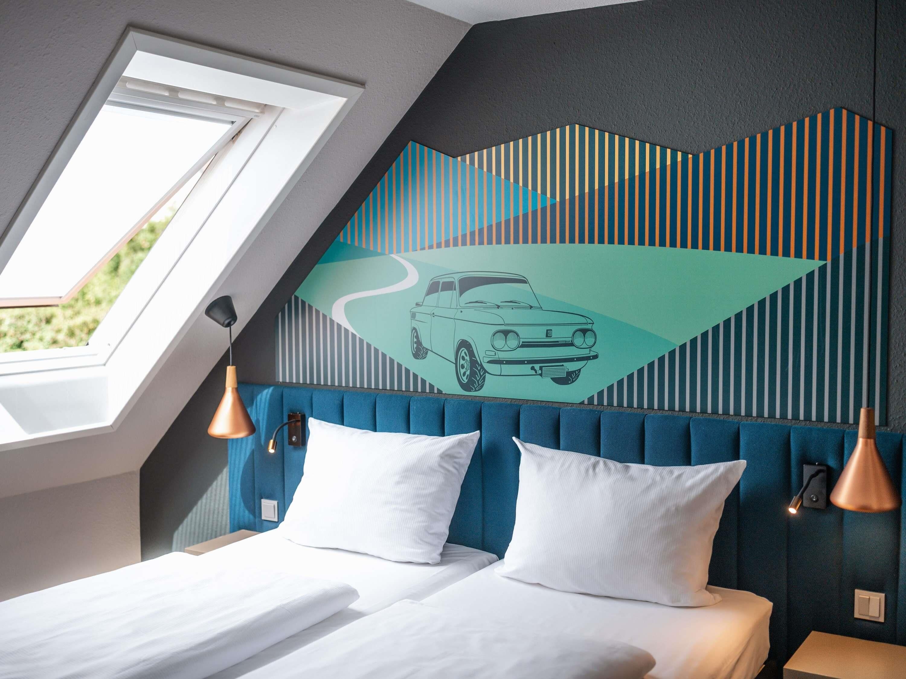 A bedroom with a sloped ceiling, a bed with white bedding, a blue upholstered headboard, and a wall mural of a car.
