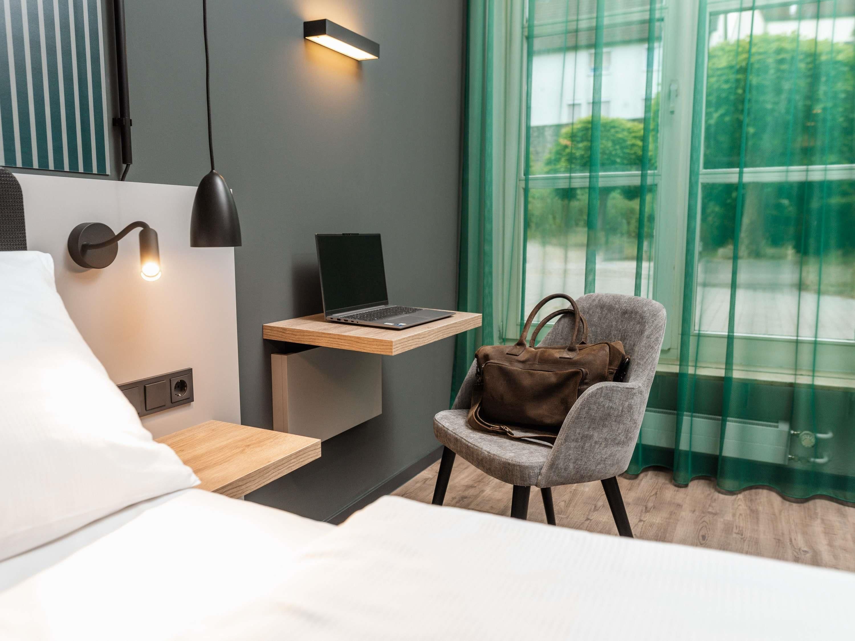 Moderni hotellihuone, jossa on sänky, työpöytä kannettavalla tietokoneella, tuoli, jossa on käsilaukku, ja ikkuna, jossa on verhot.