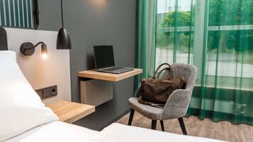 Moderni hotellihuone, jossa on sänky, työpöytä kannettavalla tietokoneella, tuoli, jossa on käsilaukku, ja ikkuna, jossa on verhot.