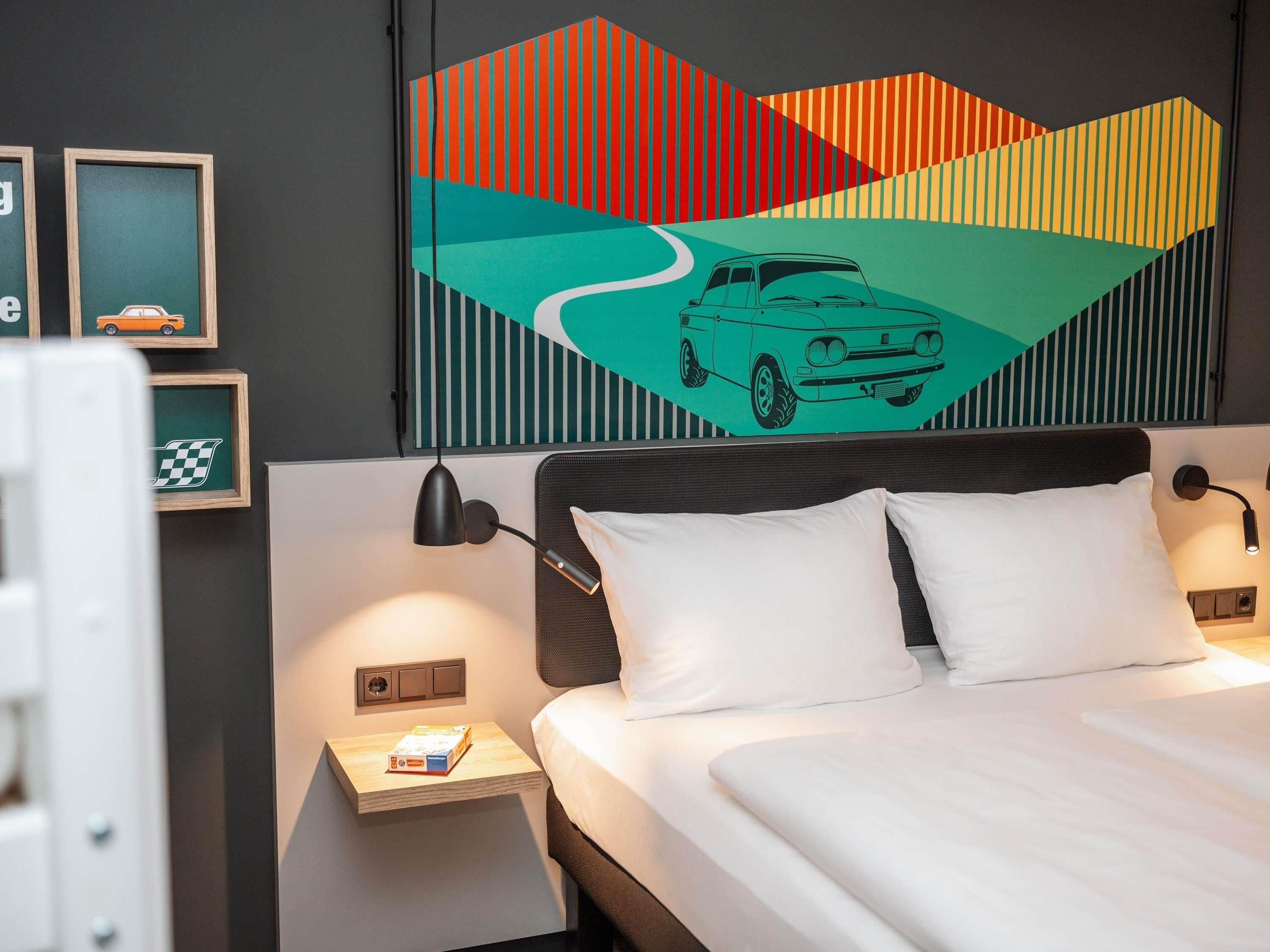 Habitación de hotel con cama, dos mesitas de noche, una lámpara de pared y un cuadro abstracto colorido sobre la cama.