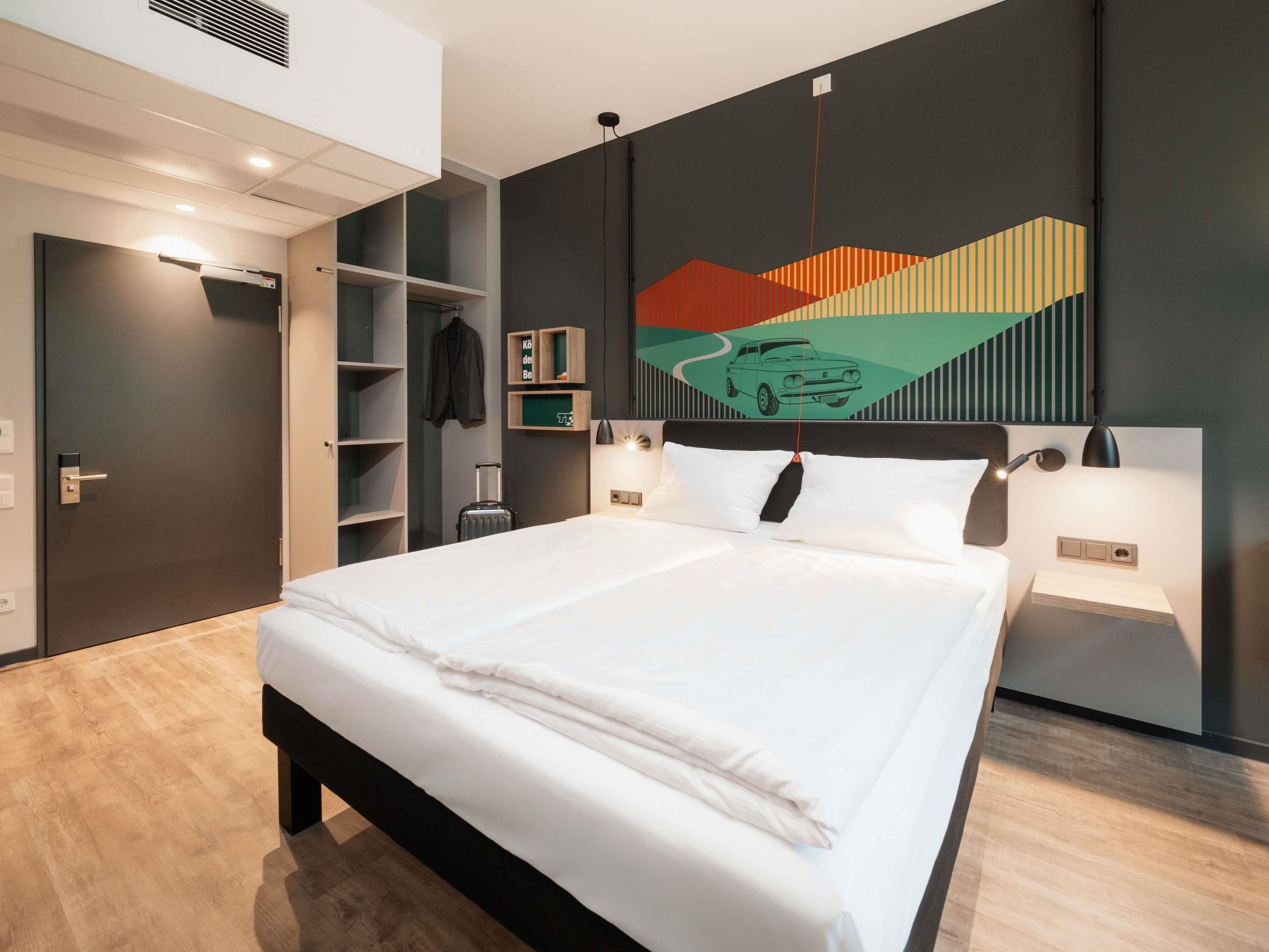 A modern hotel room with a large bed, a wardrobe, a coat rack, and a wall-mounted artwork.