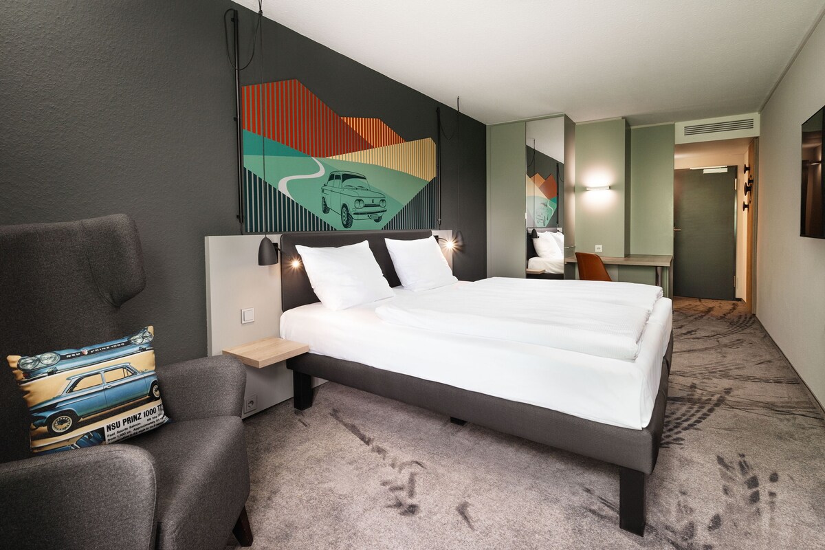 A modern hotel room with a large bed, a chair, a desk, and a wall with a colorful abstract design.