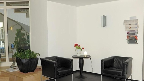 A modern room with two black leather chairs, a small round table with a vase of red flowers, a potted plant, and a staircase visible through a glass door.
