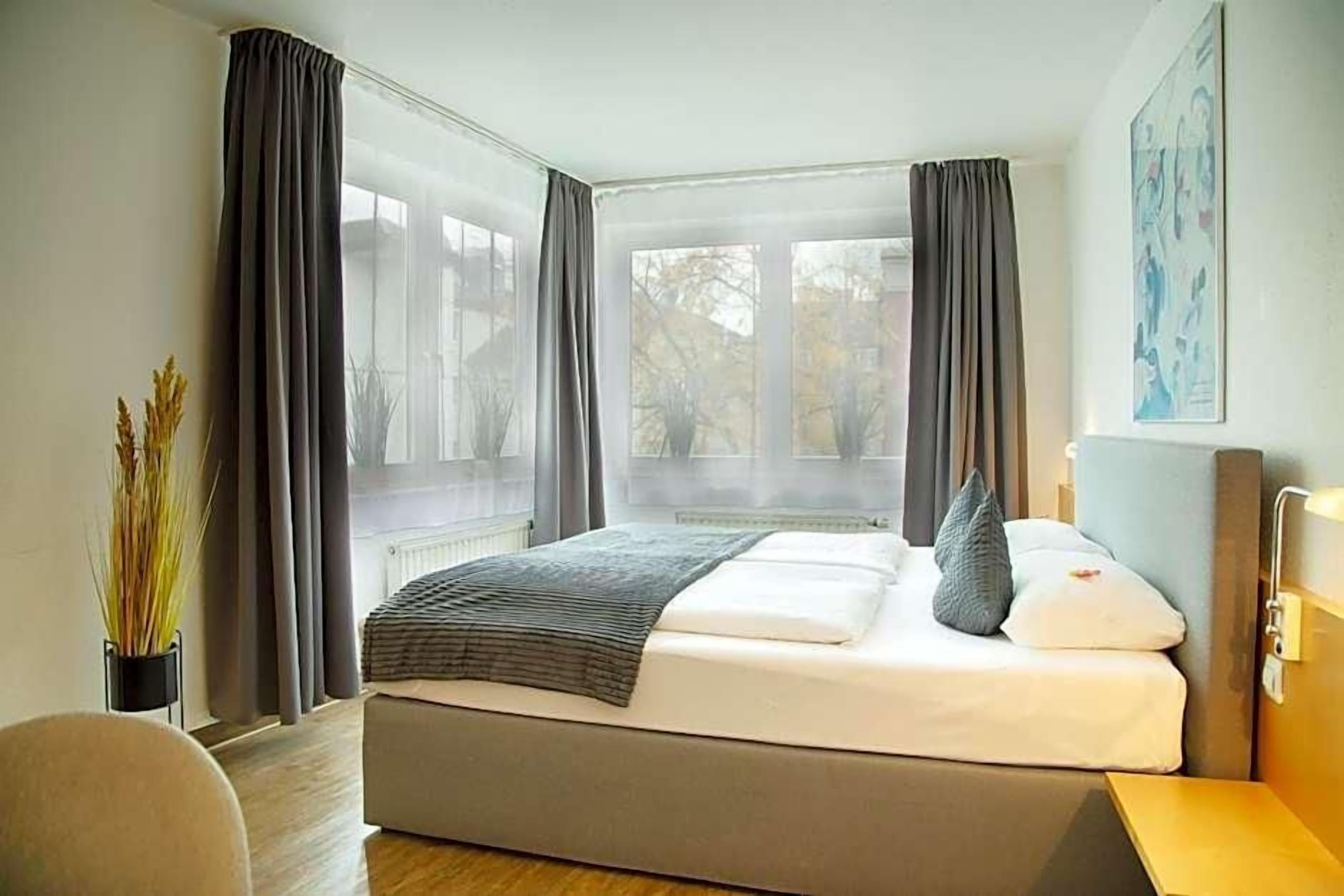 Ein modernes Hotelzimmer mit einem großen Bett, einem Nachttisch, einem Sessel und einem Fenster mit Vorhängen.