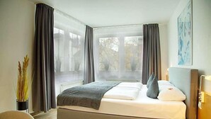 Quarto de hotel moderno com uma cama grande, uma mesa de cabeceira, uma cadeira e uma janela com cortinas.
