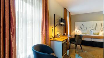 Habitación de hotel con escritorio, una silla, cama y un ventanal grande con cortinas.