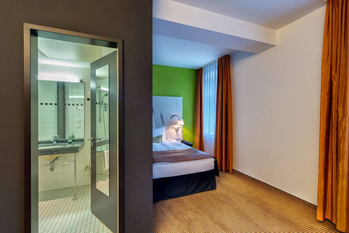 A hotel room with a bed, a bathroom with a shower, and a green accent wall.