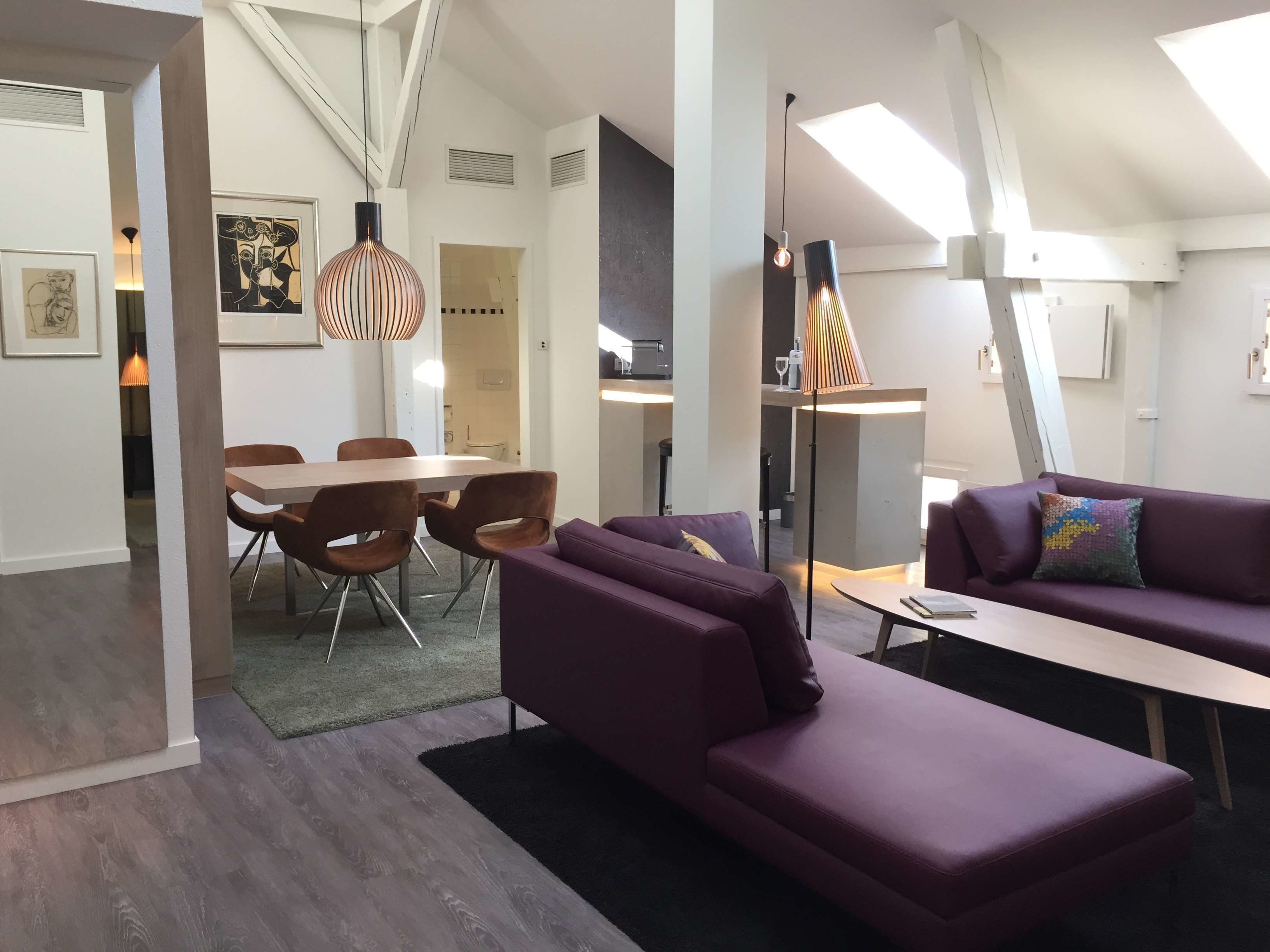 A modern living room with a purple sofa, a dining area with a wooden table and chairs, and a kitchen in the background.