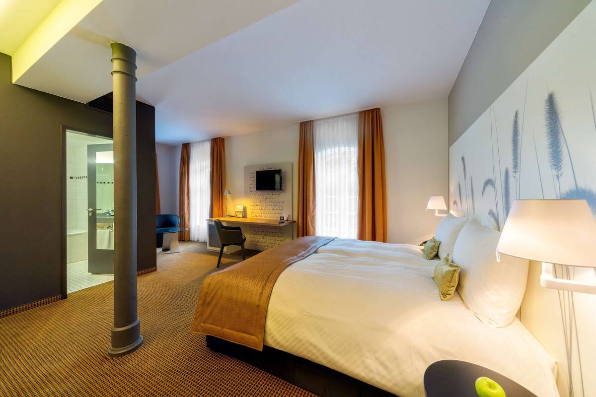 A hotel room with a bed, desk, chair, and a large window with curtains.