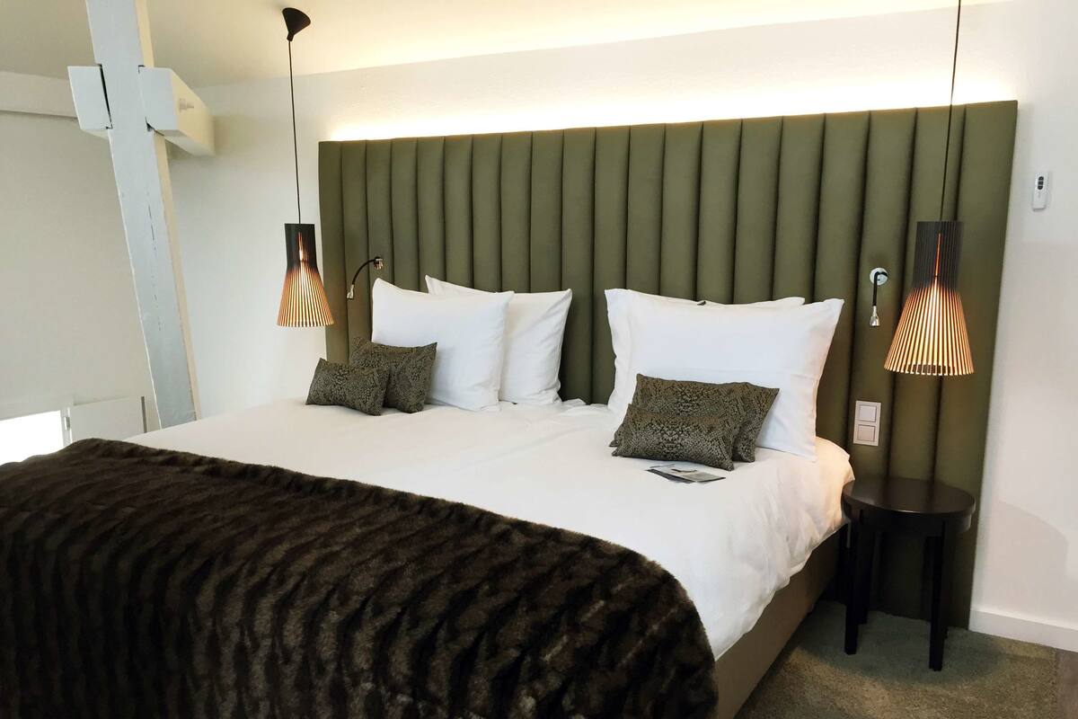 A neatly made bed with white linens and two patterned pillows, a dark throw blanket, and two bedside lamps with a unique design.