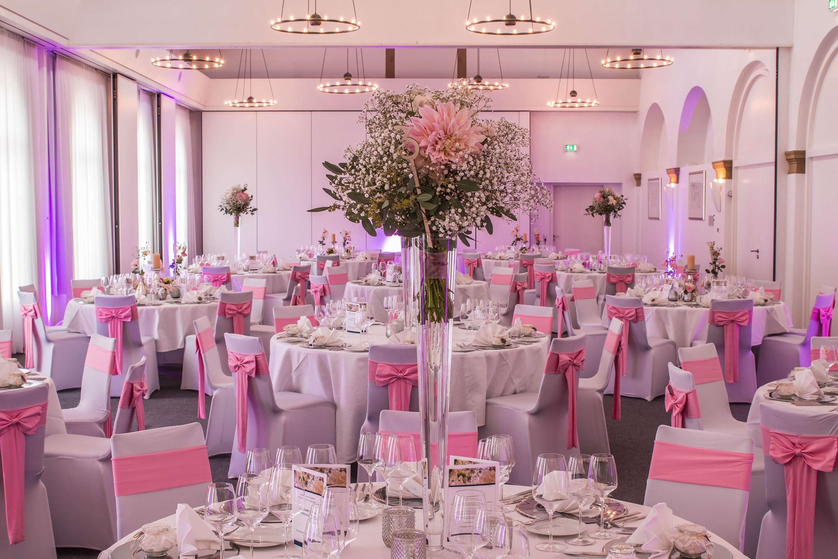 A banquet hall with round tables, pink and white tablecloths, and chairs with pink sashes.