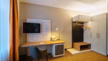 A modern hotel room with a wooden desk, a flat-screen TV, a chair, a lamp, and a wardrobe with a hanging shelf.
