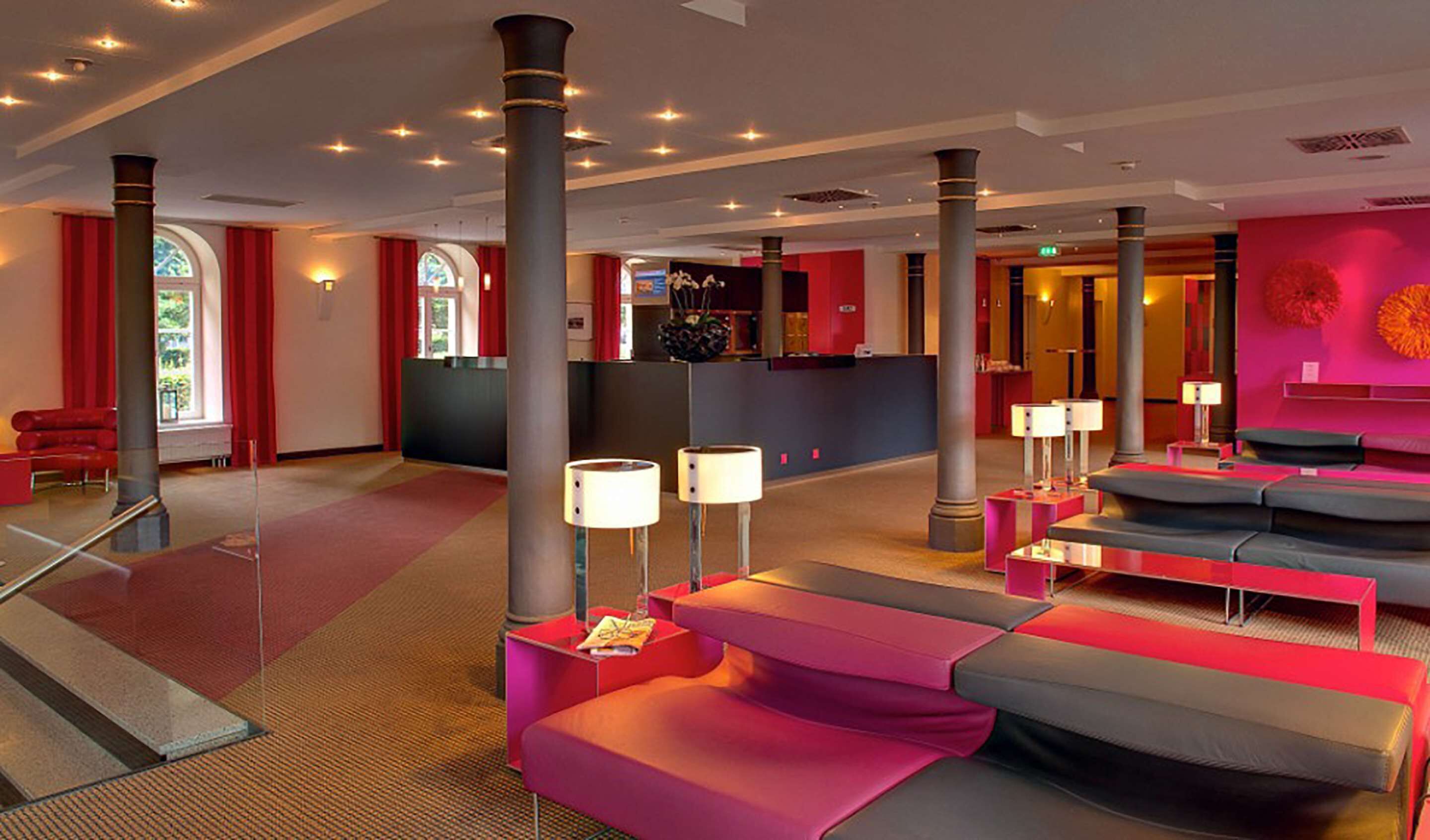 A modern hotel lobby with red and grey seating, large pillars, and a reception area.