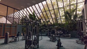 Un gimnasio con distintas máquinas de ejercicio, un ventanal con vistas a árboles y un techo con diseño de cuadrícula.