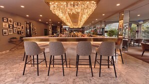 Una zona de bar con piso de mármol, taburetes de bar, una lámpara de araña grande y cuadros en la pared.