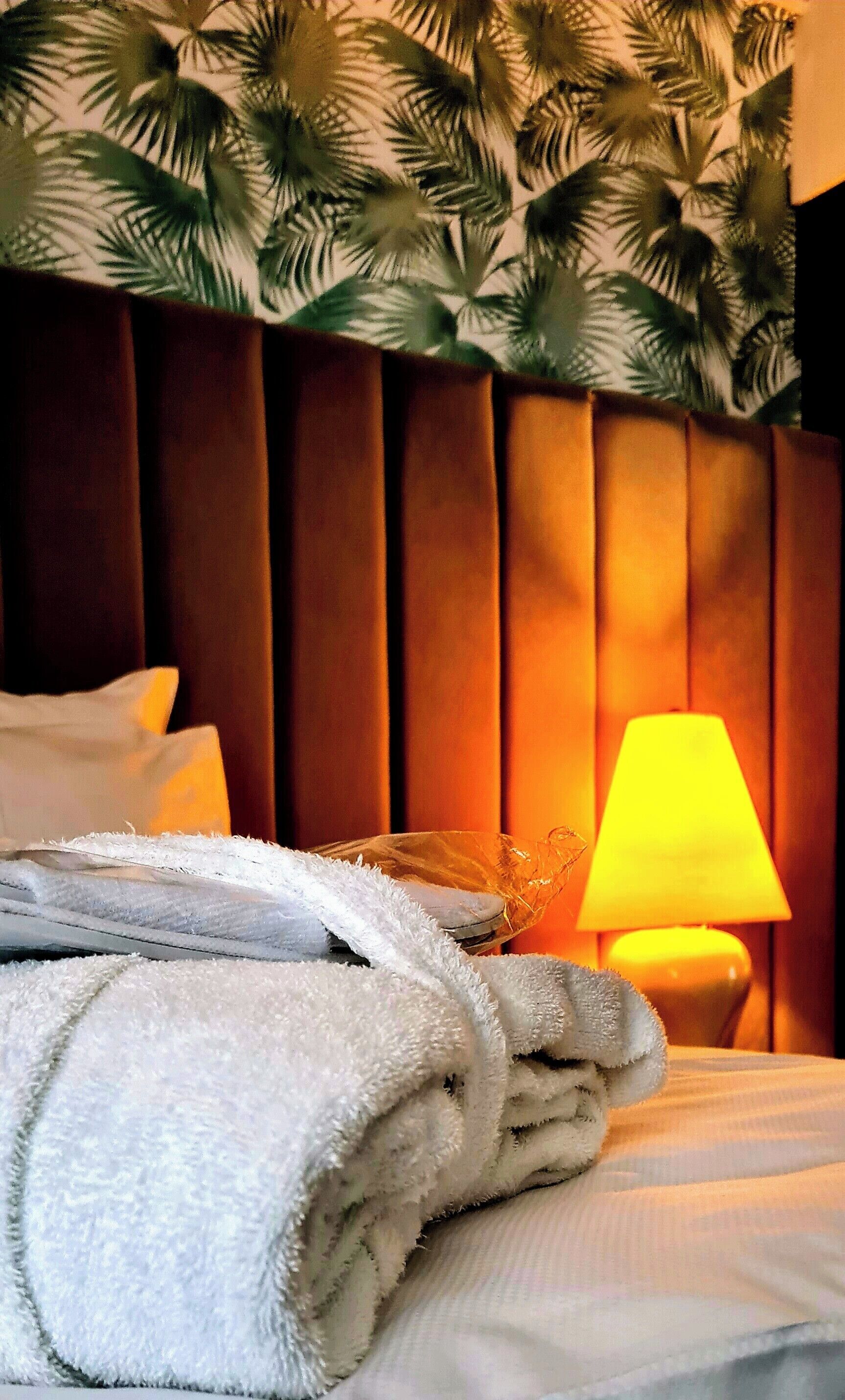 En pænt redt seng med hvide håndklæder, en lampe og en væg med bladmotiv.