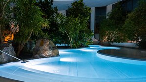 Una piscina a forma curva, circondata da piante tropicali e rocce, con un edificio moderno sullo sfondo.
