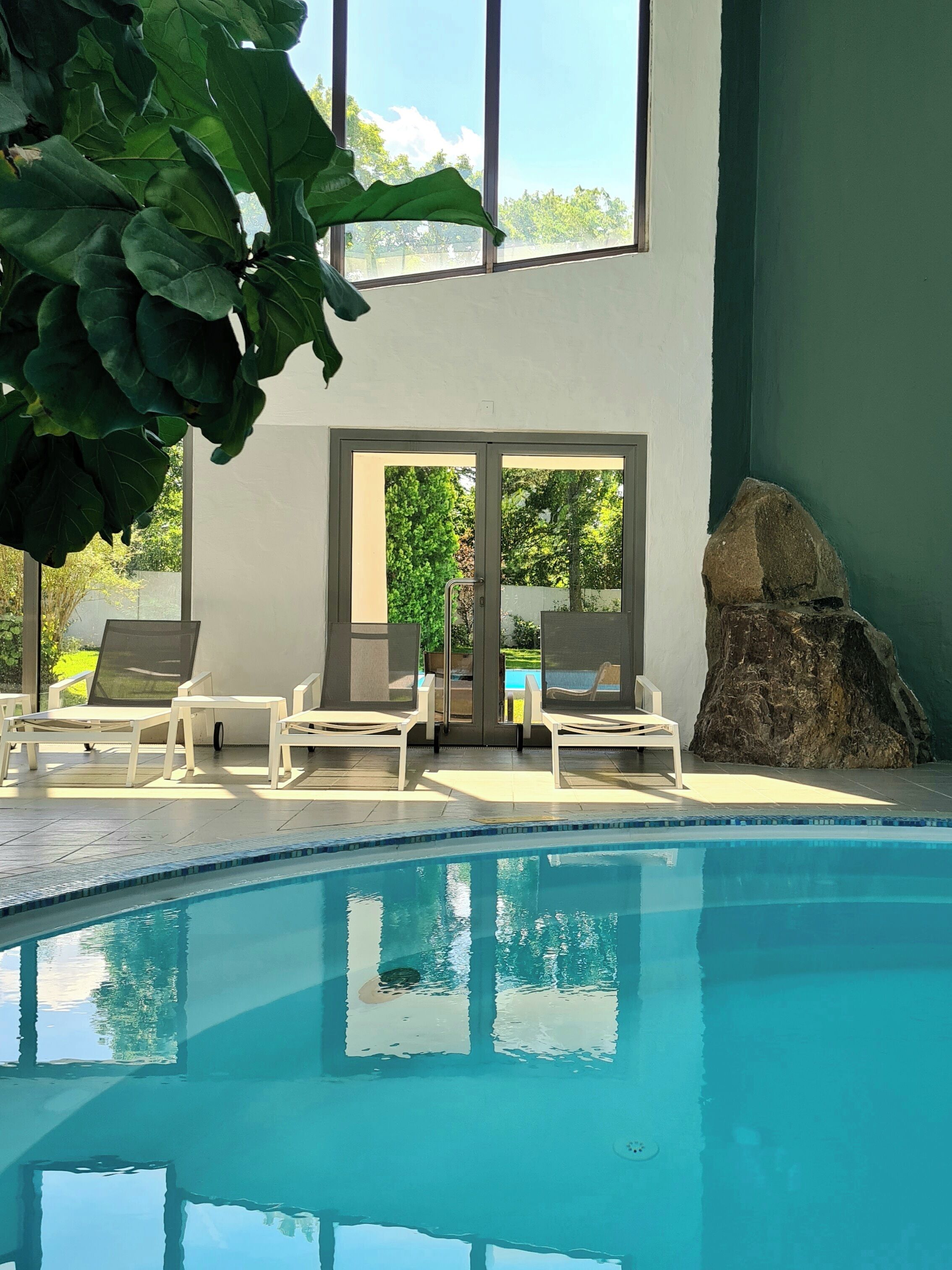 A pool area with clear blue water, white lounge chairs, and a large window overlooking greenery.