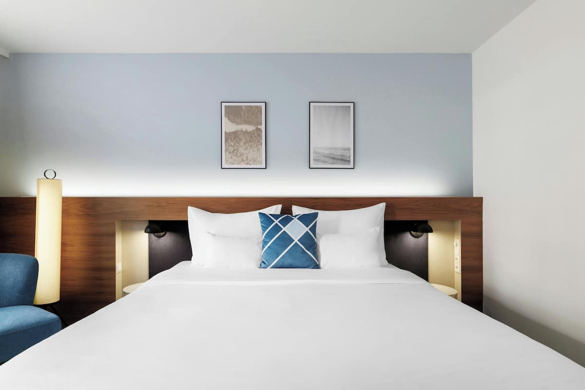 A hotel room with a bed, a blue armchair, two framed pictures on the wall, and a wooden headboard.