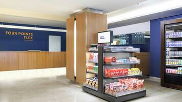 Zona de recepción de hotel con pared azul, mostrador de recepción, pantalla digital de bienvenida, exhibición de aperitivos y refrigerador para bebidas.