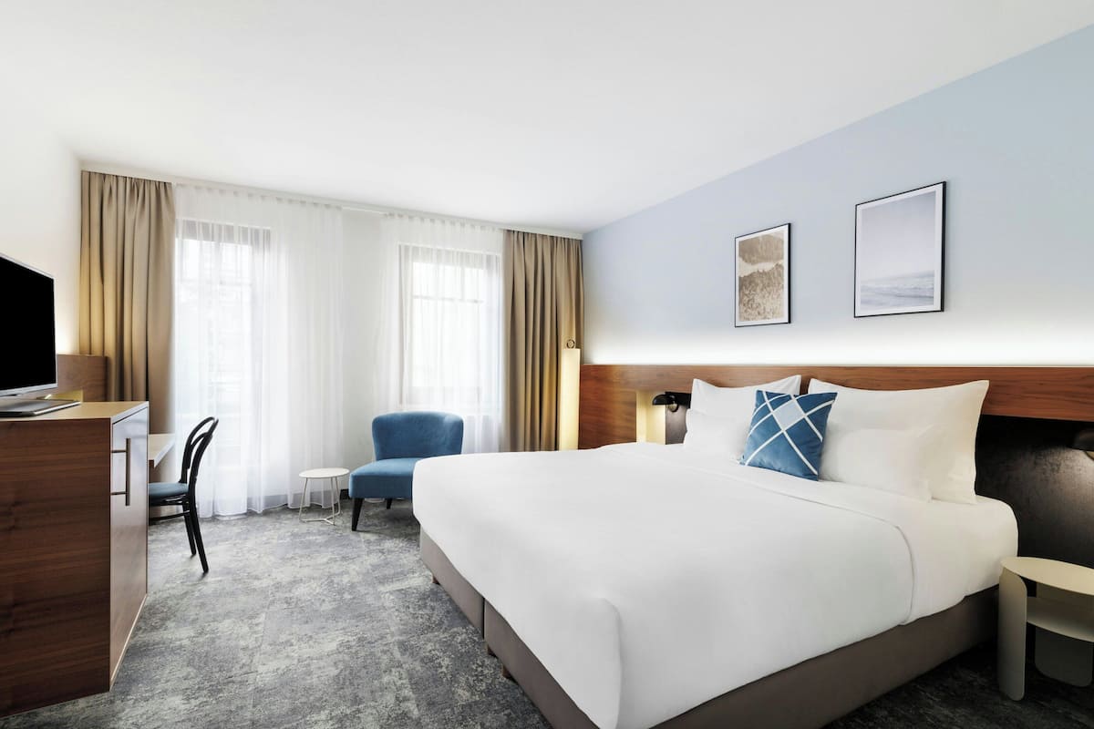 A hotel room with a bed, a desk, a chair, a small table, and a television.