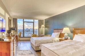 A hotel room with two beds, a desk, a chair, a TV, and a large window with a city view. - Regency Palace Amman (Amman)