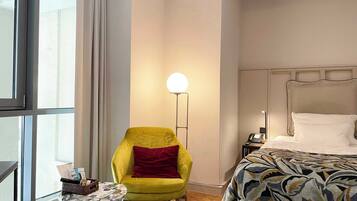 Un habitación de hotel con un ventanal grande, un sillón amarillo, una mesa redonda con una cesta, una mesita de noche con un teléfono y una colcha estampada.