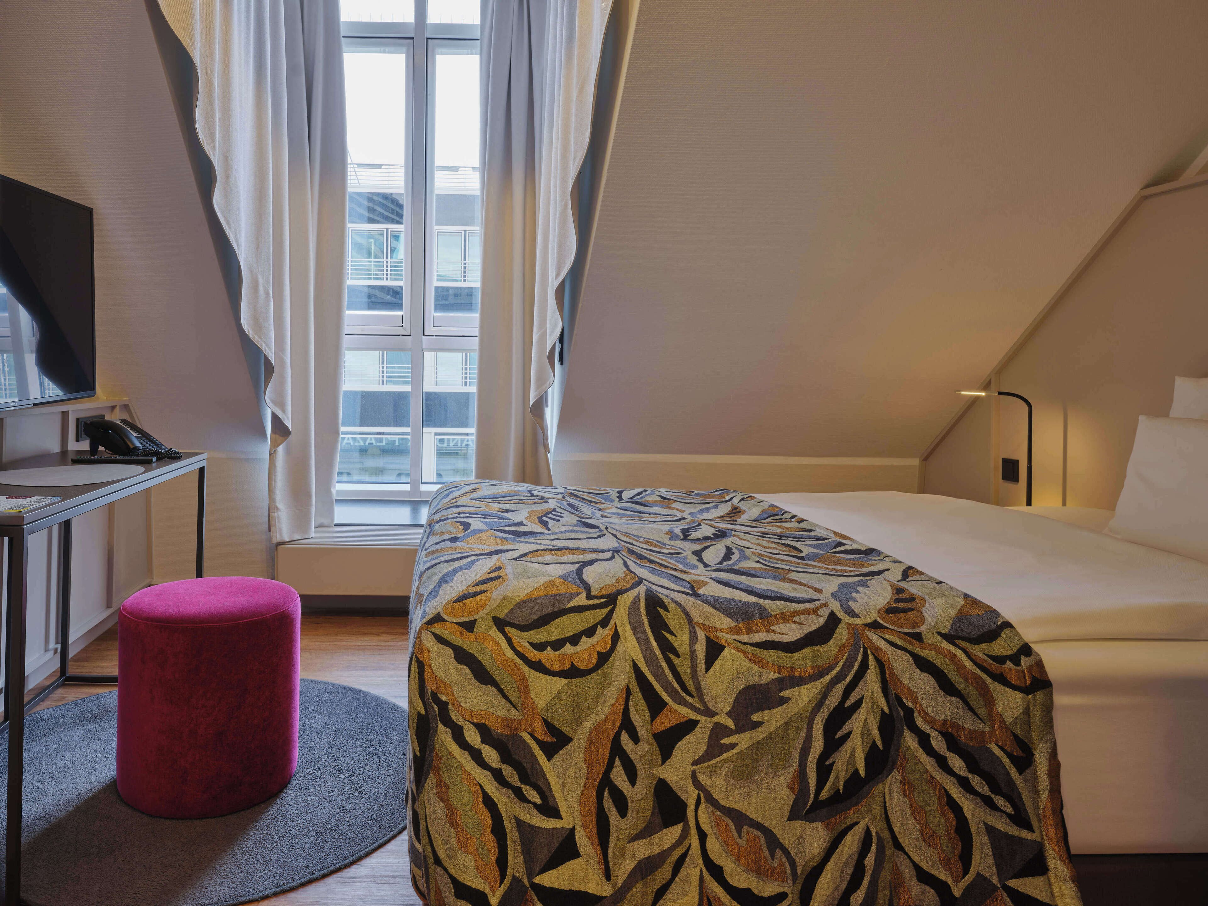 Una habitación de hotel con una cama, un escritorio con teléfono, un reposapiés rojo y una ventana con cortinas.