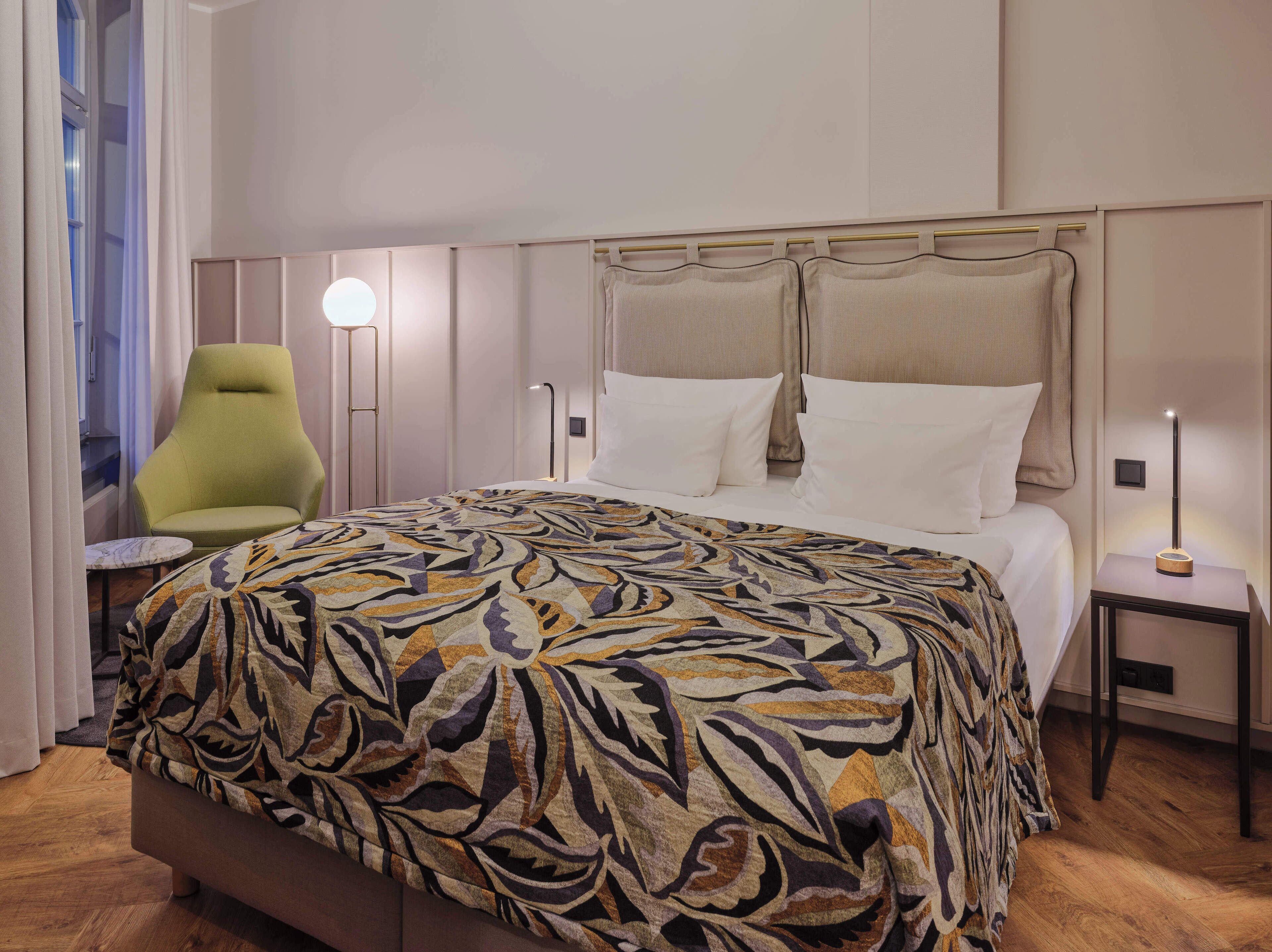 Quarto de hotel com uma cama, uma poltrona verde, uma mesa de cabeceira com luminária e uma pequena mesa também com luminária.