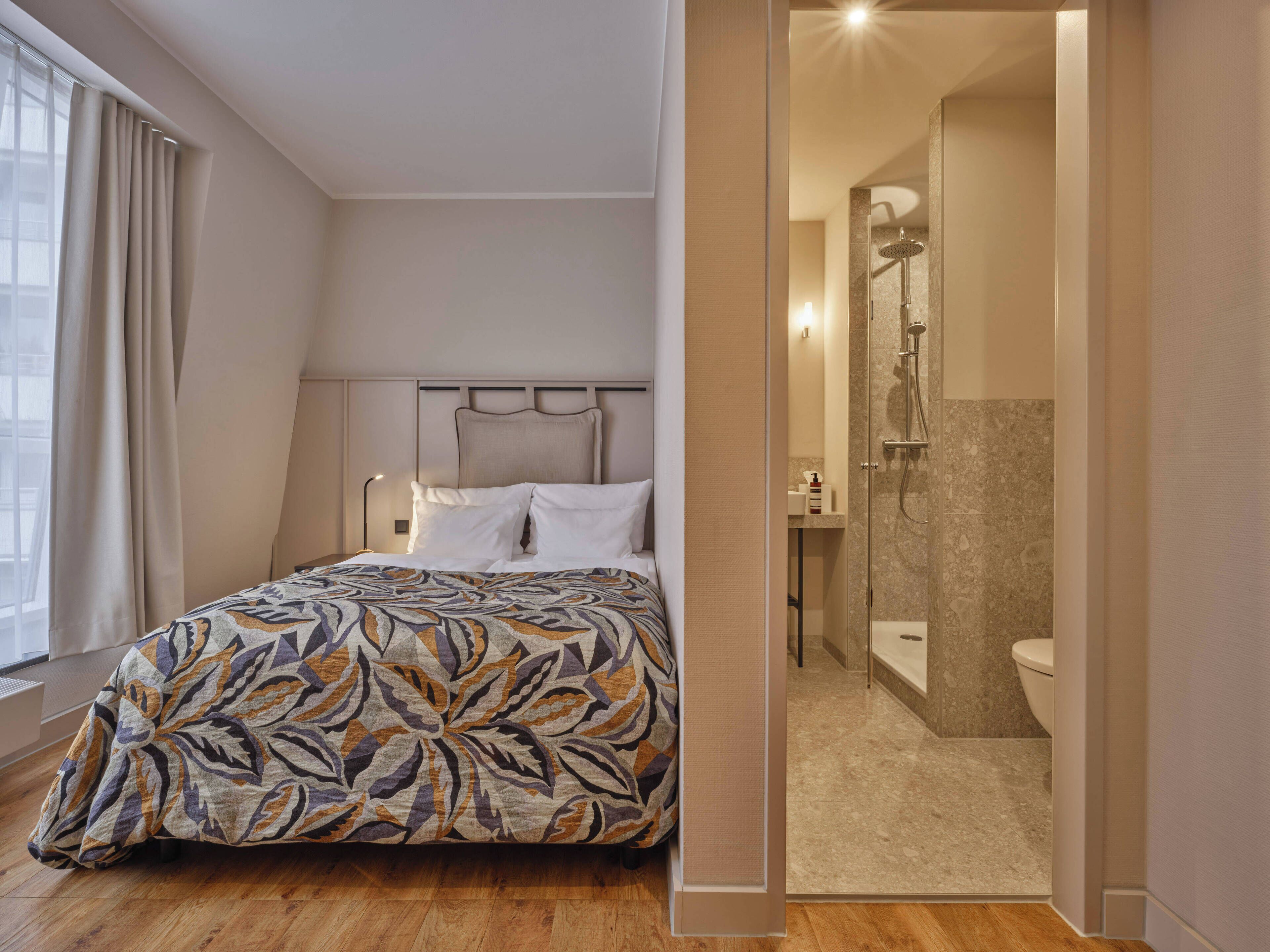 A bedroom with a patterned bedspread, a headboard, a bedside lamp, and a view of a bathroom with a shower and toilet.
