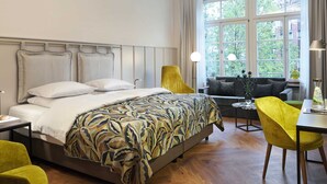 A hotel room with a bed, a sofa, a dining table, and a window with a view of trees. - Classik Hotel Alexander Plaza (Berlin)