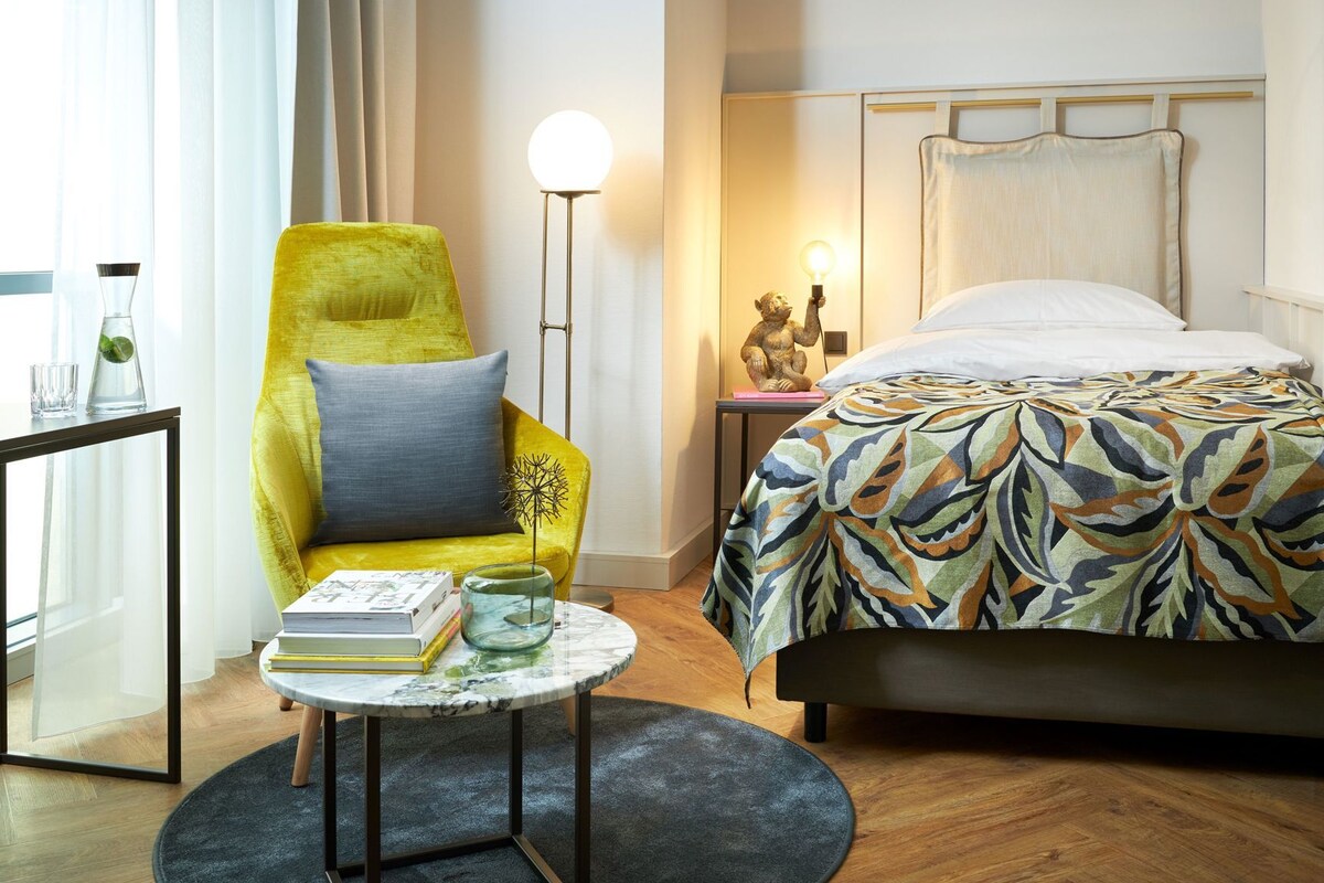 Una habitación de hotel con una cama, un sillón amarillo, una mesa redonda con libros, una lámpara de pie y una mesita de noche con una estatua.