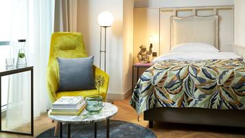 A hotel room with a bed, a yellow armchair, a small table with books and a vase, and a floor lamp.