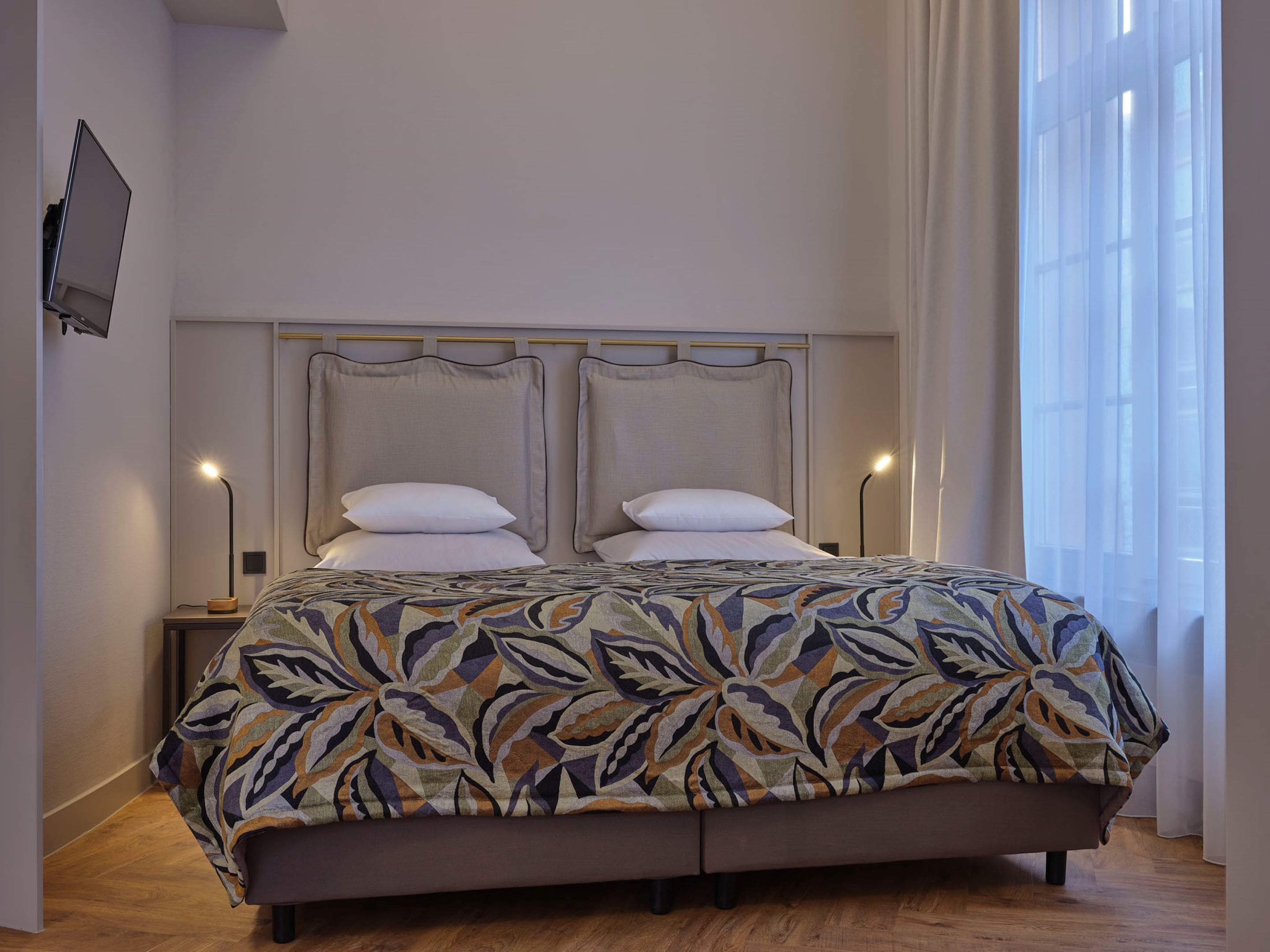 Ein Doppelbett mit einem gemusterten Bettbezug, zwei Nachttische mit Lampen, ein Flachbildfernseher an der Wand und ein Fenster mit durchsichtigen Vorhängen.