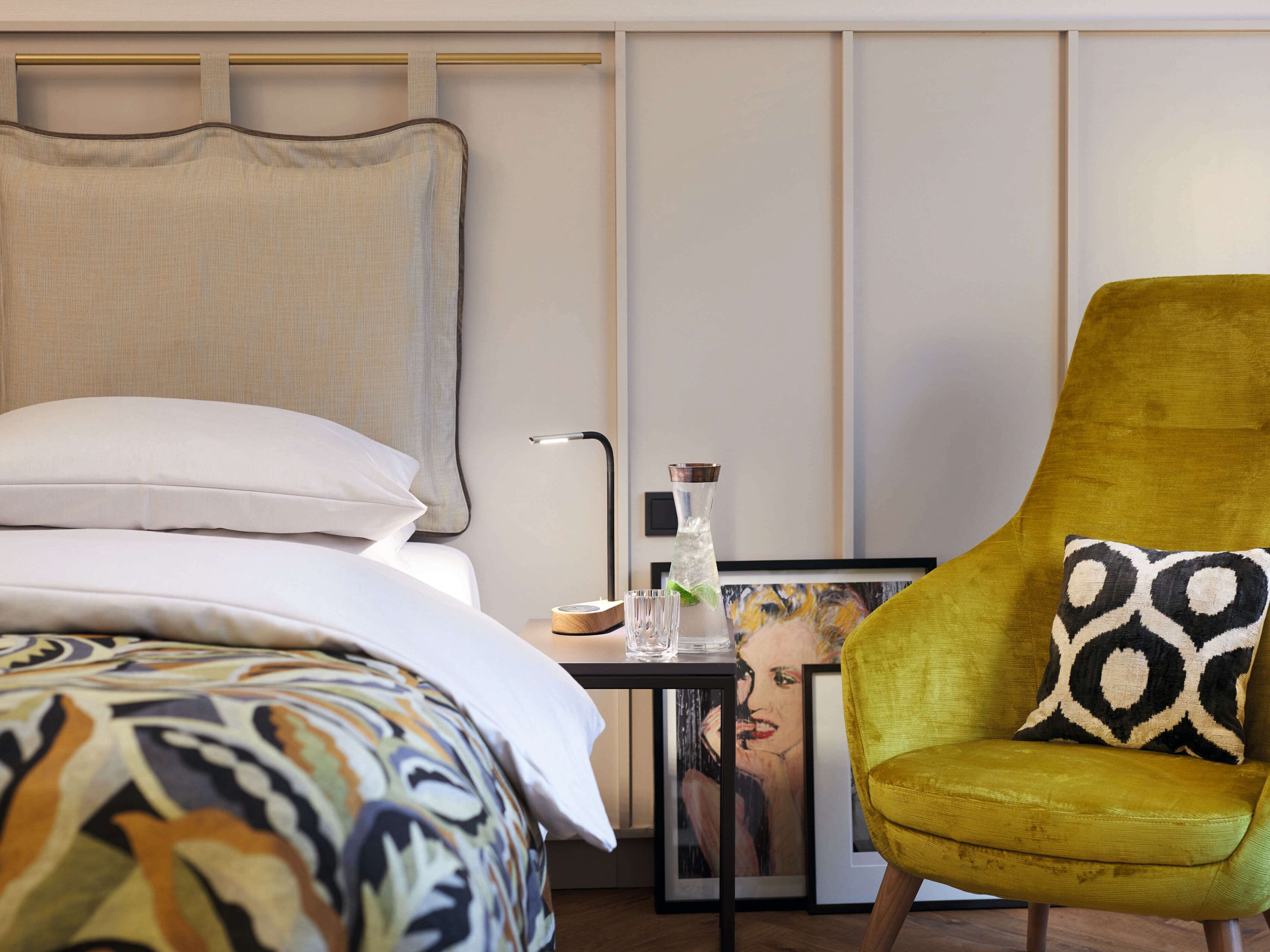 Ein modernes Hotelzimmer mit einem Bett, einem Nachttisch mit Lampe, einem Sessel und einem gerahmten Bild an der Wand.