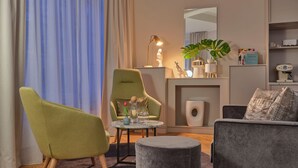A modern living room with a green armchair, a grey sofa, and a round ottoman. There is a small table with a vase and a lamp. A plant is on a shelf, and a coffee machine is on the counter. - Classik Hotel Alexander Plaza (Berlin)