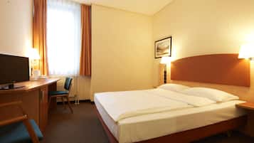 Ein Hotelzimmer mit einem Bett, einem Schreibtisch mit Fernseher, einem Sessel und einem Fenster mit Vorhängen.
