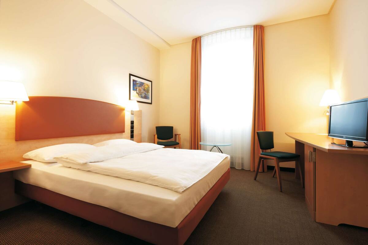 Une chambre d’hôtel avec un lit, deux chaises, un bureau avec une télévision et une fenêtre avec des rideaux.