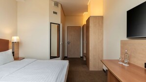 Une chambre d’hôtel comprenant un lit, une lampe de chevet, un téléviseur à écran plat fixé au mur, un bureau en bois avec une bouteille et un verre, et une porte donnant sur une autre pièce.