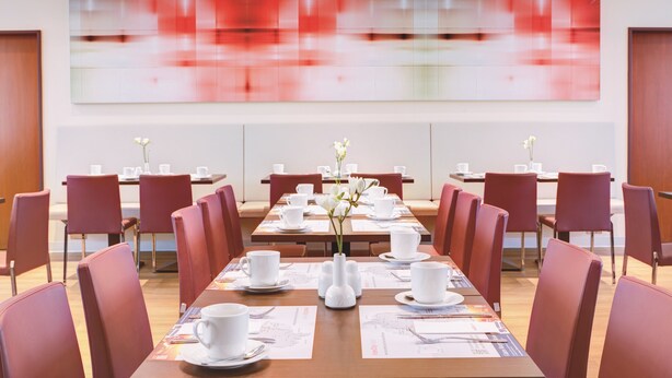 Een vergaderruimte met lange tafels, rode stoelen, wit servies en een groot abstract schilderij aan de muur.