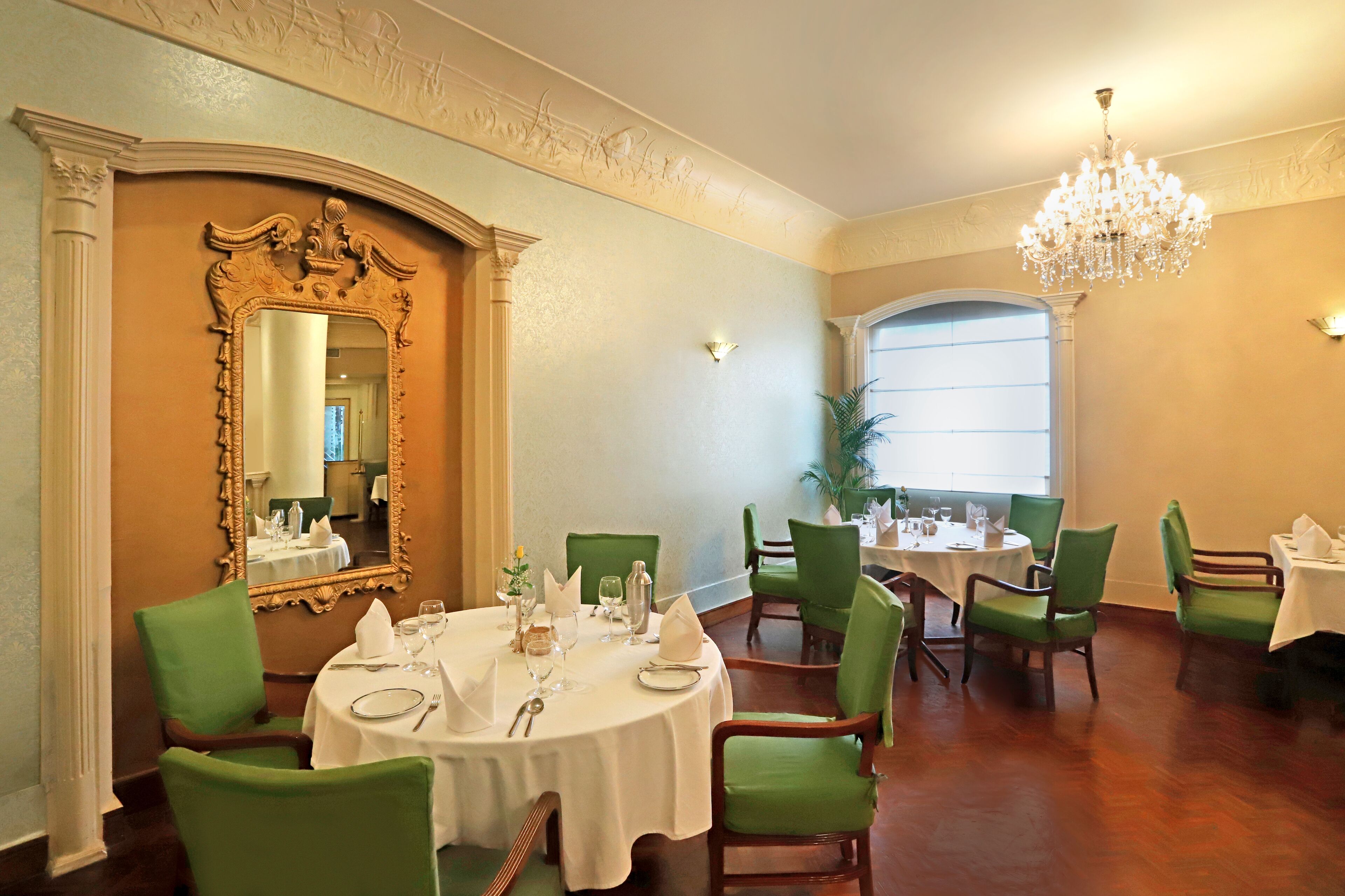 A dining area with round tables, green upholstered chairs, a large ornate mirror, and a chandelier.