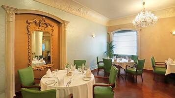 A dining area with round tables, green upholstered chairs, a large ornate mirror, and a chandelier.