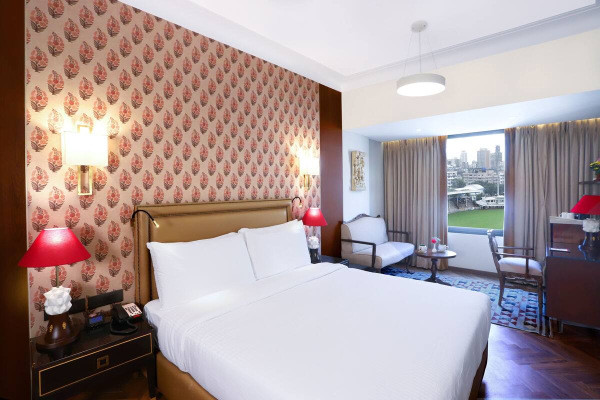 Un dormitorio de hotel con una cama grande, dos lámparas de noche, un escritorio con una silla, vistas de la ciudad a través de la ventana y un papel tapiz estampado.
