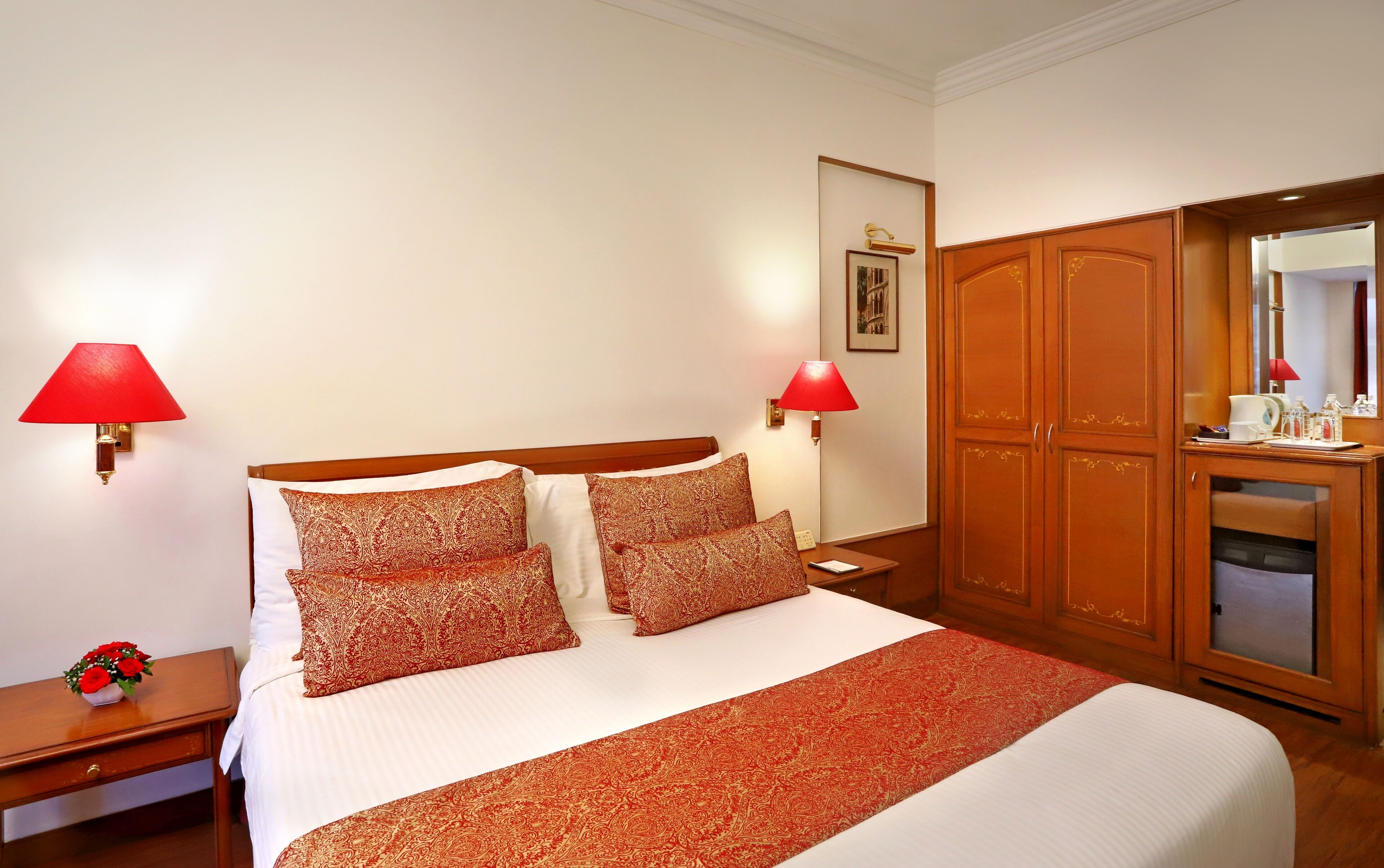 A hotel room with a large bed, two bedside lamps, a wooden wardrobe, and a small table with a vase of flowers.