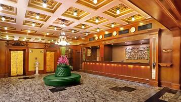 Zona de recepción de hotel con suelo de mármol, una maceta decorativa verde con flores, una lámpara de araña y un mostrador de recepción con un reloj y una escultura en la pared.