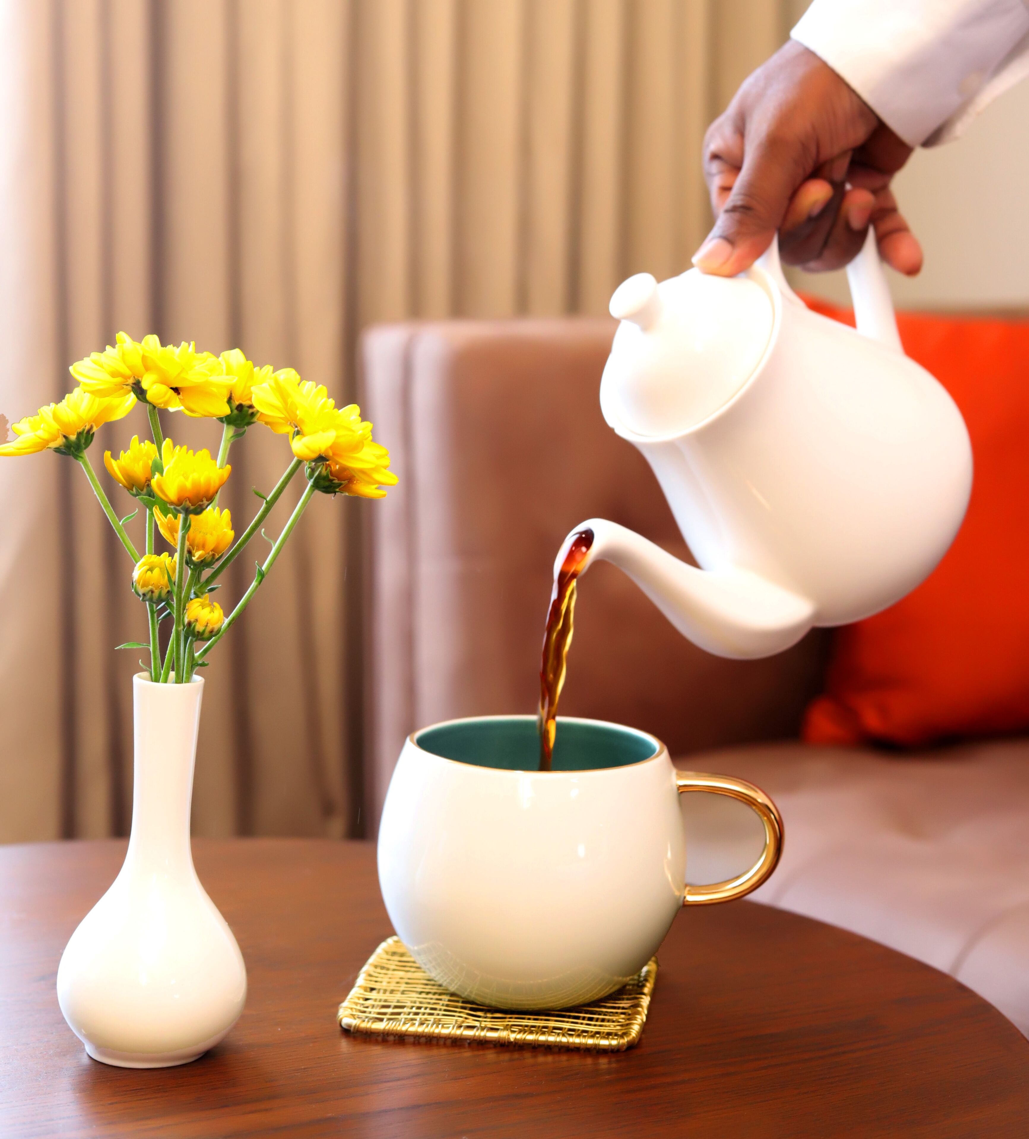 Una tetera blanca vertiendo té en una taza blanca con asa dorada, sobre una mesa redonda de madera.