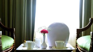 Una mesa de comedor con una bandeja que contiene una jarra blanca, una taza blanca, un platillo blanco y una tetera blanca. En el centro, hay un florero blanco con flores rosadas. Dos sillas de madera con cojines verdes están situadas a ambos lados de la mesa.