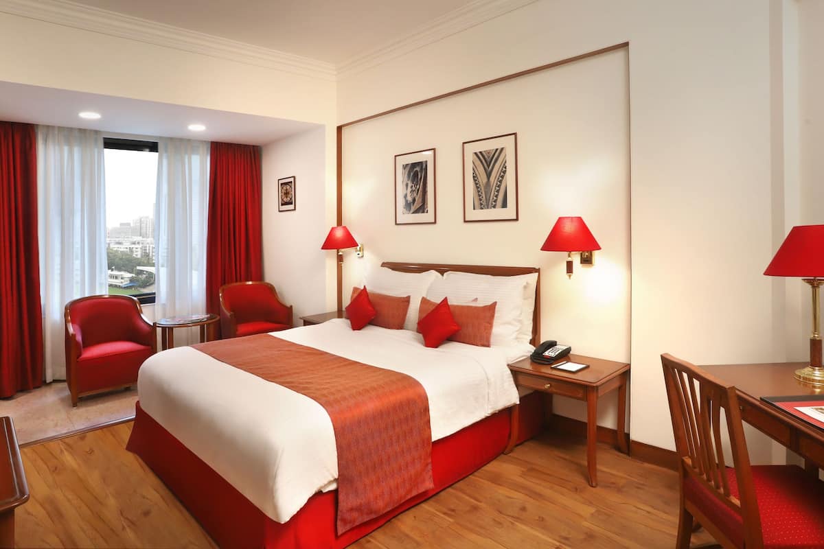 Habitación de hotel con una cama grande, dos sillas rojas, un escritorio y vista a la ciudad.