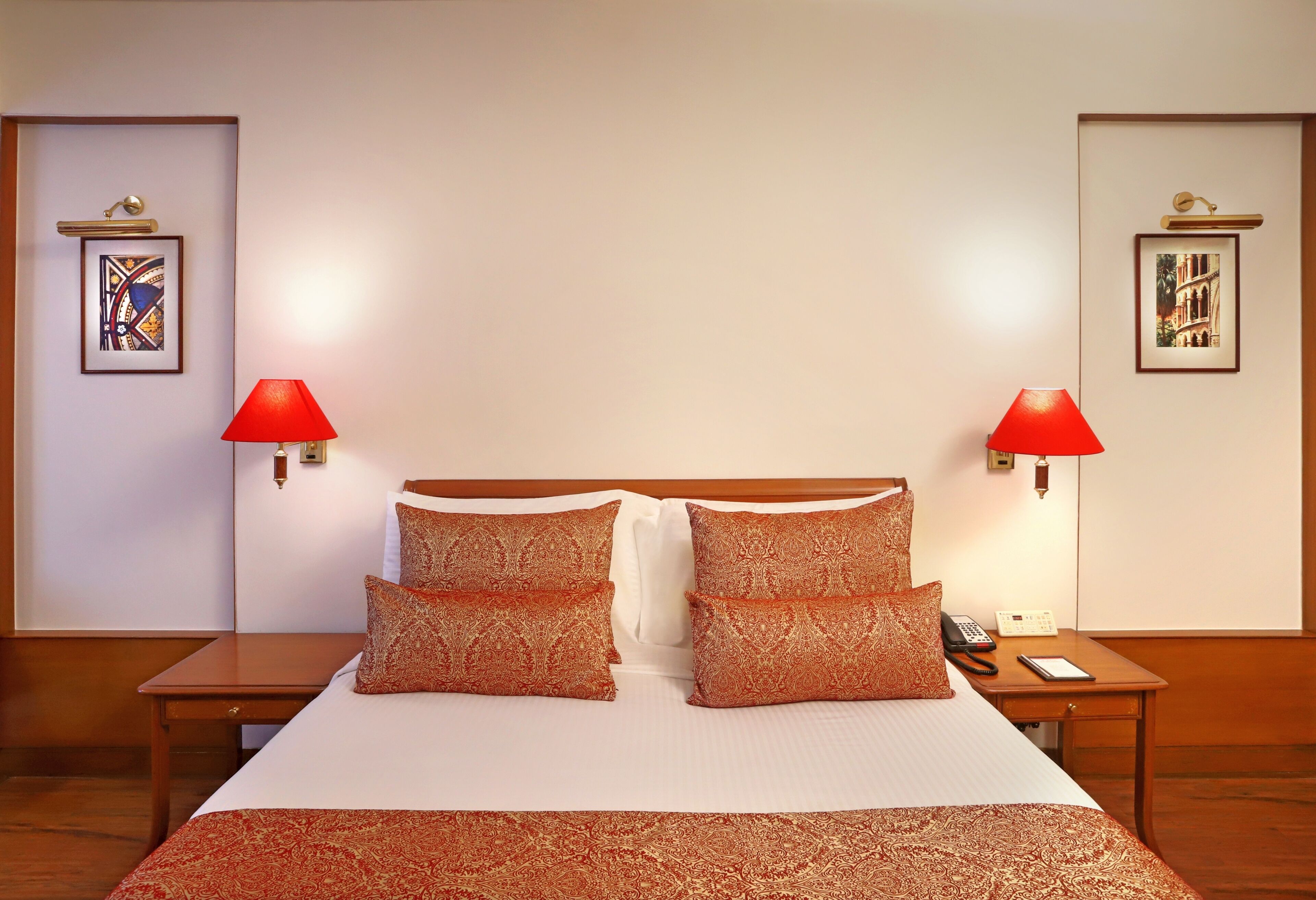 A hotel room with a large bed, two bedside tables, red lamps, and framed pictures on the walls.